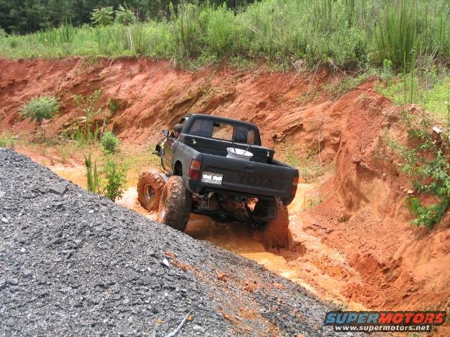 Black Toyota pickup truck with large tires driving through a muddy ditch on a dirt trail