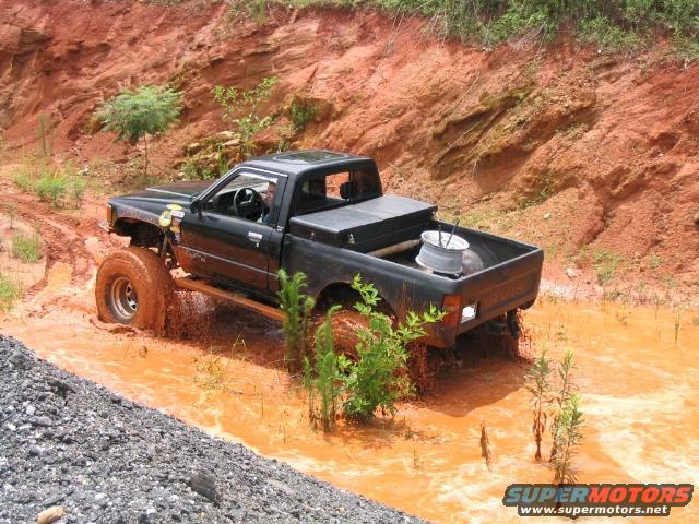 Black pickup truck with large tires driving through mud, aftermarket toolbox in bed