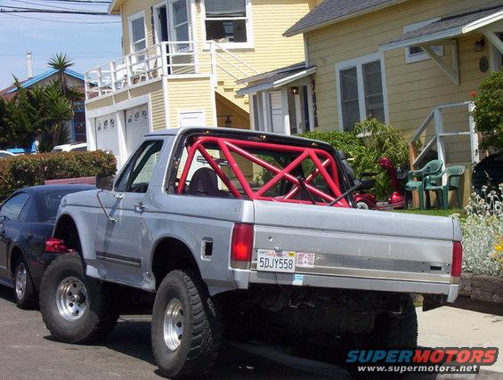 Silver Ford Bronco with red roll cage, lifted, parked next to a dark car