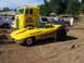 1994 Ford F-150 - A modified yellow 1965 Chevrolet Corvette convertible is parked in a dirt lot with a large yellow truck and other vehicles.