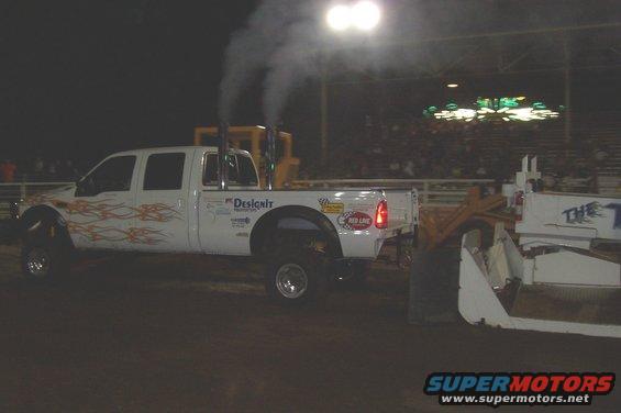 White pickup truck with flame decals, dual exhaust stacks, and "Designit" text on the side, pulling a large white implement. Crowd in background