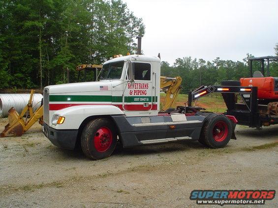White semi-truck with red and green stripes, carrying a mini-excavator on a trailer