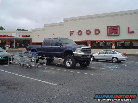 Lifted blue Ford F-250 truck parked in a grocery store lot. Silver sedan in background