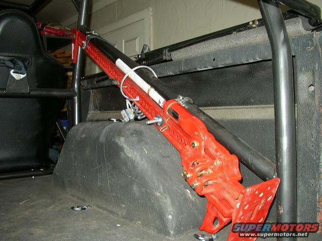 Red hi-lift jack secured with straps inside a vehicle, near a black seat and roll cage