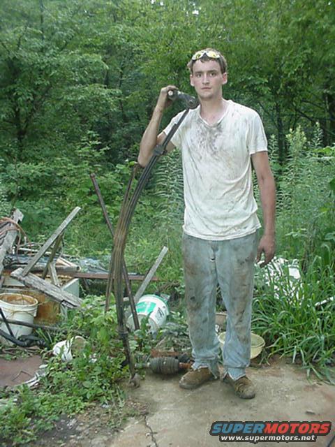 Man holding a large, curved metal car part in a cluttered outdoor setting