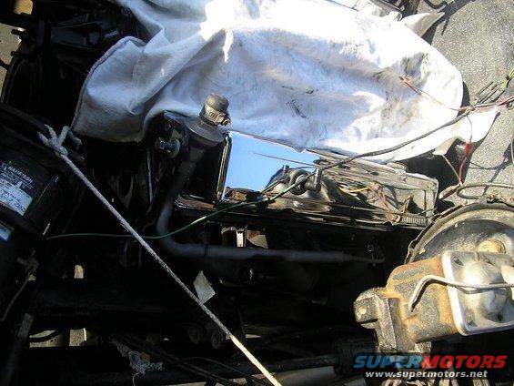 Engine bay detail showing a chrome valve cover, part of a brake master cylinder, and some wiring. A white cloth covers part of the engine