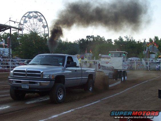 Silver Dodge Ram pickup truck with black smoke billowing from exhaust, pulling a sled at a tractor pull event. Ferris wheel in background