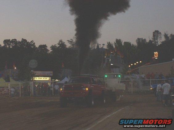 Red pickup truck pulling a trailer, emitting a large cloud of black smoke. Fairground in background