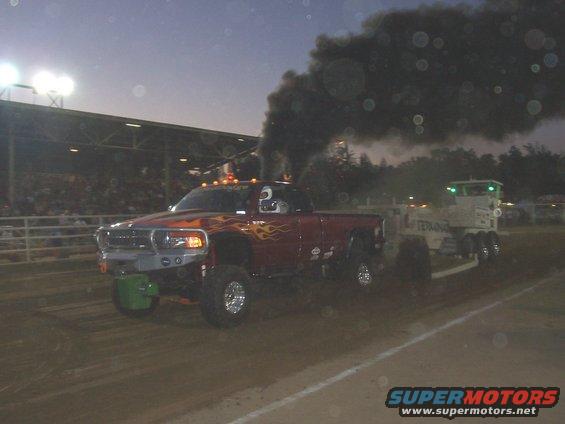 Red pickup truck with flame decals and massive exhaust smoke, pulling a trailer at an event