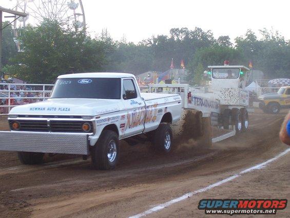 White Ford pickup truck with a large chrome bumper pulling a trailer labeled TERMINATOR. Another yellow truck is visible in the background