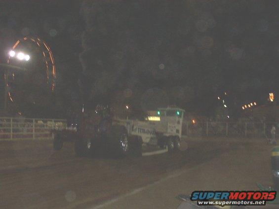 Tractor pull truck with large rear tires on a dirt track at night, Ferris wheel in background