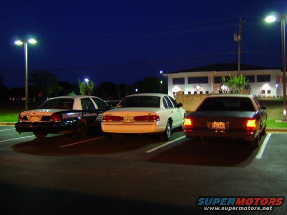 Three sedans parked in a lot at night. The middle car is white, the left car is a police car, and the right car is dark colored