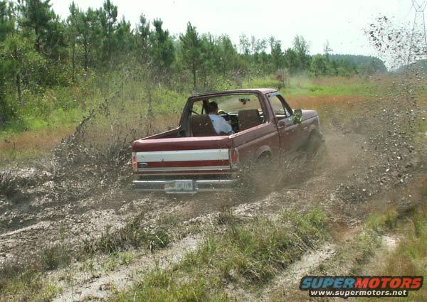 Red 1988 Ford Bronco driving through a muddy field, kicking up water