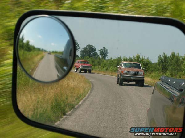 Brown truck with rusty grille and white roof, seen from side mirror. Another red truck is ahead