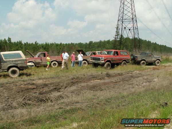 Several trucks and a red Bronco parked in a muddy area with a power line tower in the background