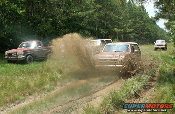 Mud-splattered 1987 Ford Bronco driving through a muddy track with trees in the background