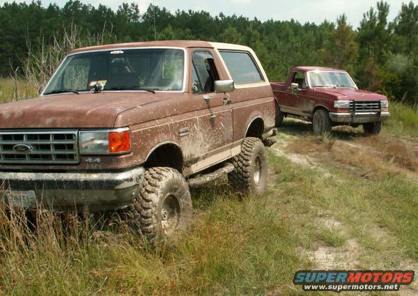 Brown Ford Bronco and red Ford pickup truck on a dirt path with trees in the background
