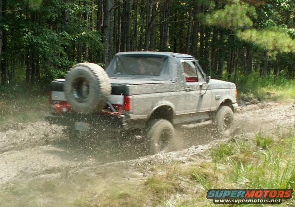 Mud-splattered Ford Bronco driving through mud, spare tire mounted on rear