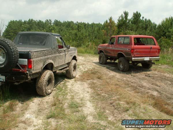 Muddy 1980s Ford Bronco with soft top and spare tire on rear, next to a red 1987 Ford Bronco on a dirt track