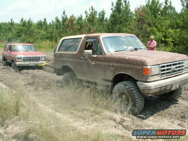 Two Ford Broncos stuck in mud, one brown and one red. A person stands in the background