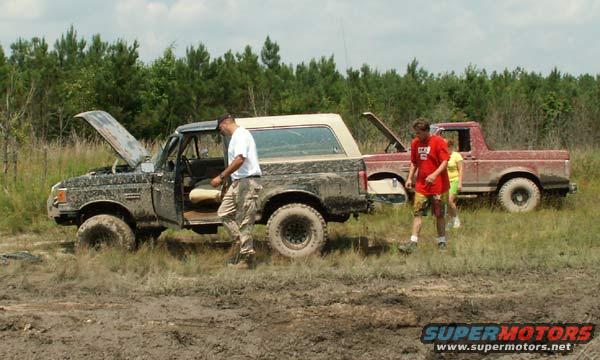 Mud-covered black Ford Bronco with hood open, driver's door ajar, in a muddy field