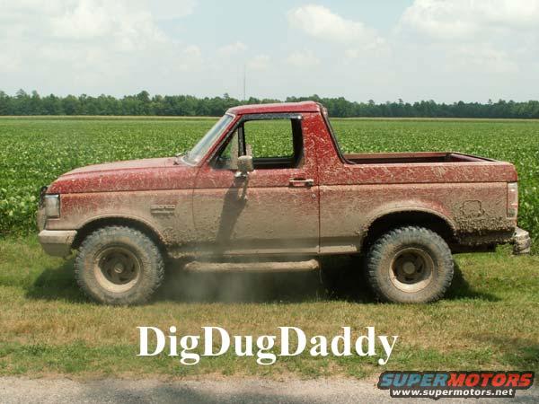 Mud-covered red Ford Bronco parked on grass with a field behind it