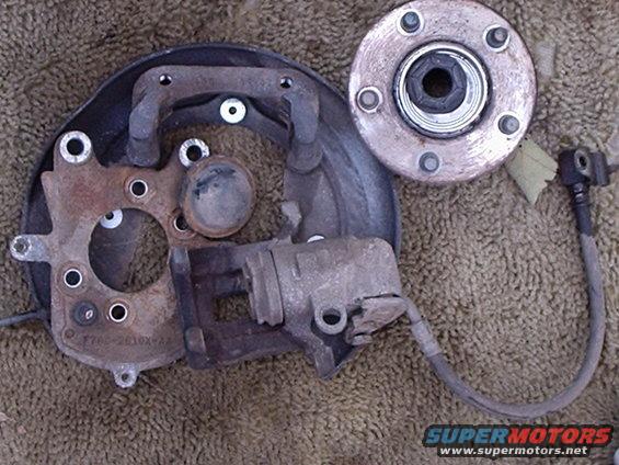 Car brake caliper, rotor, and hub assembly on a textured surface. Part number visible on bracket