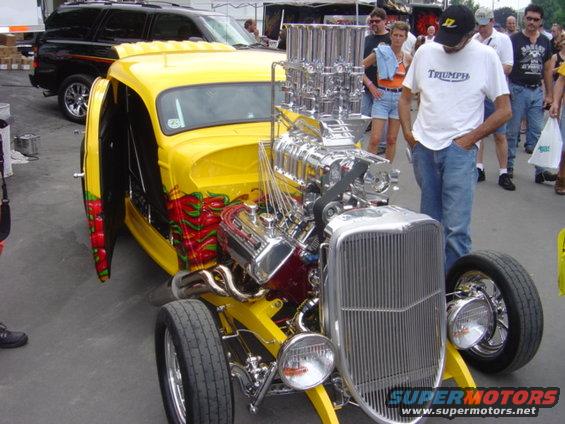 Yellow hot rod with a massive supercharger and exposed engine, parked next to a black SUV
