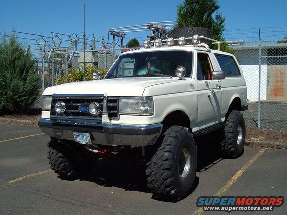 White 1989 Ford Bronco lifted with large off-road tires, roof rack, and extra lights