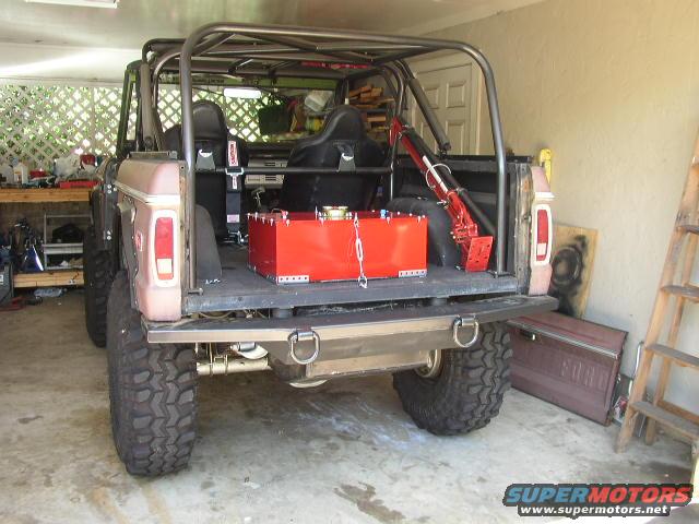 Rear view of a 1978 Ford Bronco with roll cage and red fuel tank. A detached Ford door leans against the wall