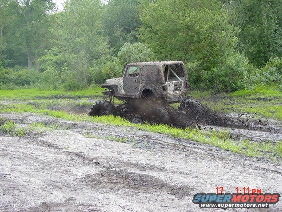 Modified Jeep stuck in mud, tires deep in muck, trees and grass in background. Watermark reads SUPERMOTORS