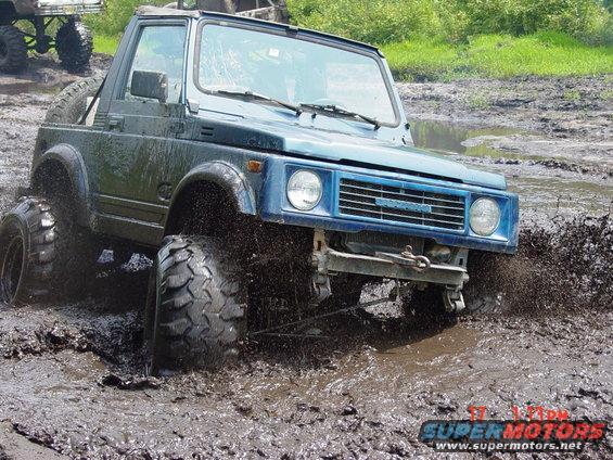 Blue Suzuki Samurai off-roading in mud, front passenger side tire deep in muck. Another vehicle is visible in the background