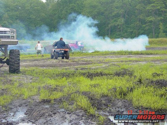 ATV kicking up smoke with people and trucks in muddy field