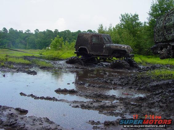 Mud-covered off-road vehicle stuck in mud with large tires. Another vehicle is partially visible to the right