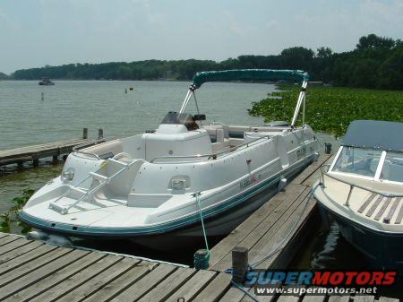 Pontoon boat docked next to a smaller boat on a lake with lily pads and trees in the background