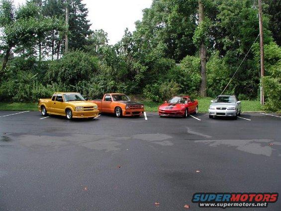 Yellow pickup truck, orange pickup truck, red sports car, and silver sedan parked in a lot
