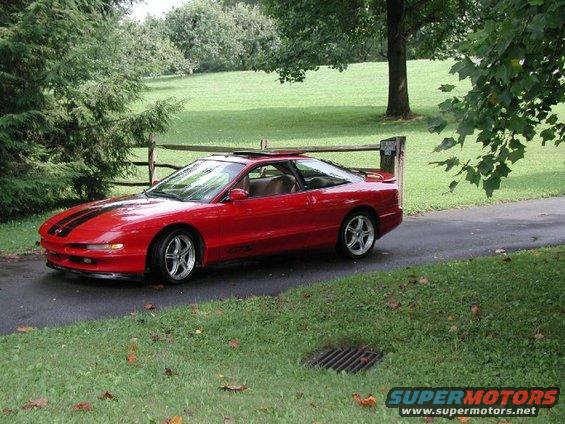 Red sports car with black racing stripes on the hood, parked on a paved driveway next to a wooden fence
