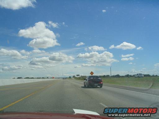 Dark red 1994 Ford Probe driving on a highway, rear view. Another white vehicle is parked on the shoulder