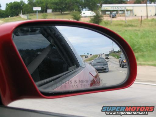 Traffic on a highway reflected in a red car's side mirror. Text on mirror reads 'Objects in mirror are closer than they appear'