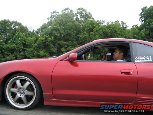 Red coupe side view, driver holding camera, aftermarket wheels, 'Vision' sticker on window