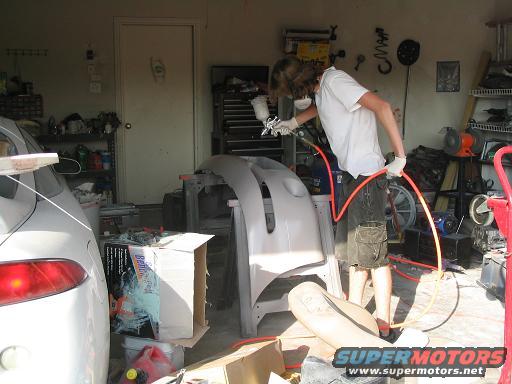 White car bumper on stand, being spray painted by a person in a garage
