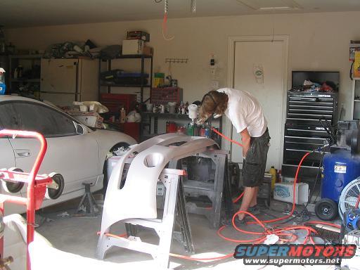 Person painting a car bumper in a garage with a white car in the background