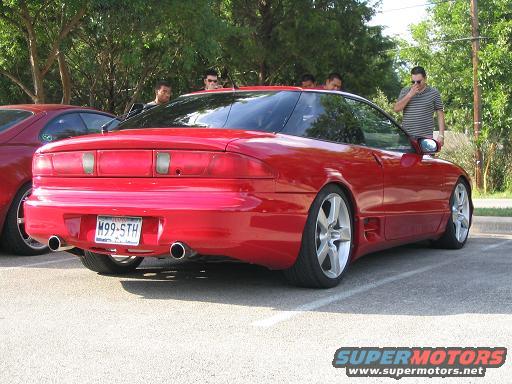 red sports coupe with custom wheels and dual exhaust, another red car to the left, people standing around