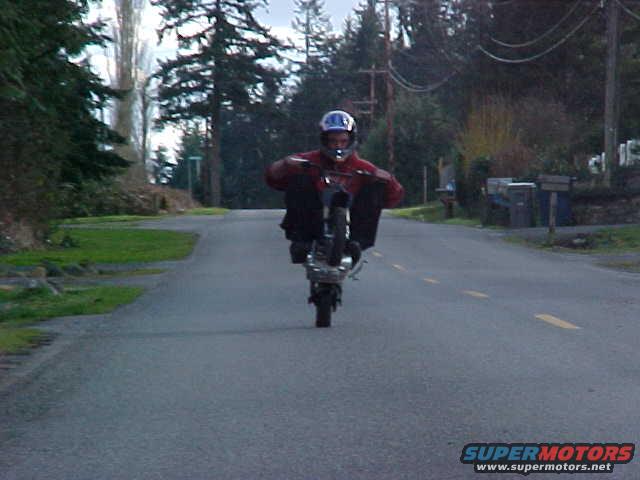 Person on a dirt bike performing a wheelie on a paved road with trees and houses in the background. Watermark reads SUPERMOTORS.NET