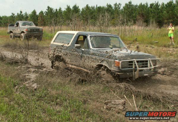 Mud-splattered Ford Bronco driving through a muddy field with another truck behind it