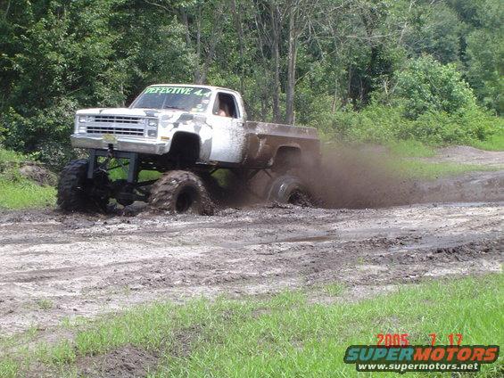 White pickup truck with camouflage decals on the hood, lifted with large tires, driving through mud