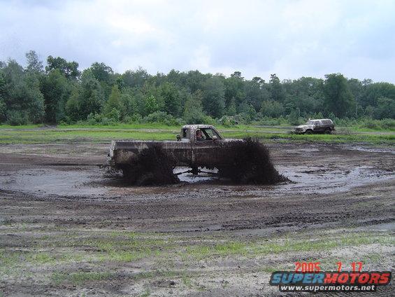 Mud-splattered pickup truck driving through a muddy field, side view. Another SUV is visible in the background
