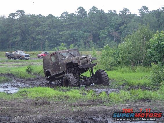 Mud-covered Jeep Wrangler with oversized tires stuck in mud, two trucks in background