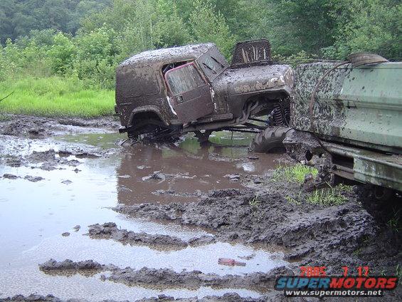 Two off-road vehicles stuck deep in mud, one is a brown Jeep-like vehicle with its door open, the other is a green truck