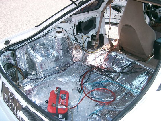 Car interior stripped down, showing sound deadening material on floor and rear panels, with battery and wiring visible